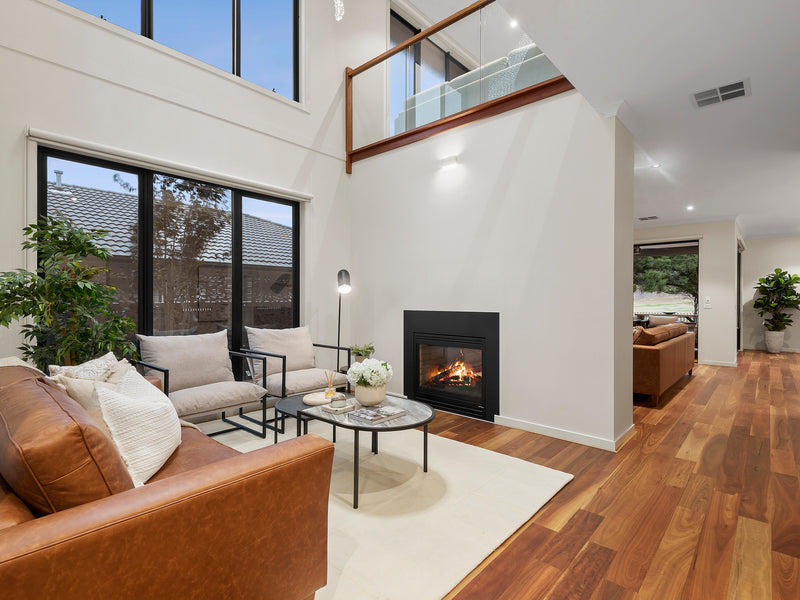 Double-height living room home staging featuring leather sofa, neutral armchairs, fireplace and timber flooring, styled to highlight space, warmth and buyer appeal.