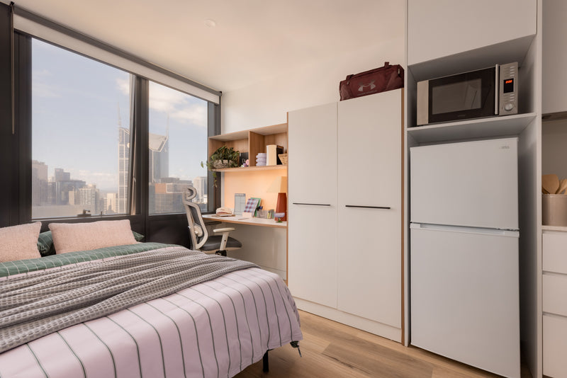 Small bedroom with bed, desk, and kitchenette area with refrigerator and microwave.